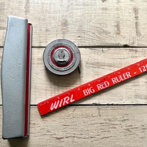 Puede incluir: Una colección de herramientas de medición vintage, que incluye una caja de cinta métrica plateada, una cinta métrica circular y una regla roja. La regla está marcada como "WIRL BIG RED RULER - 1290". La caja de la cinta métrica está marcada como "MODEL NO. 3".