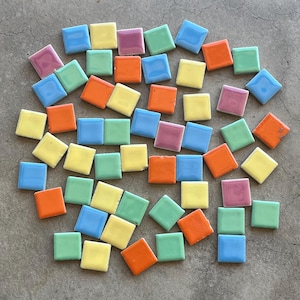 Könnte beinhalten: Eine Sammlung kleiner, quadratischer Keramikfliesen in verschiedenen Pastellfarben, darunter Gelb, Blau, Orange, Grün und Rosa. Die Fliesen sind auf einer grauen Oberfläche verstreut und bieten eine lebendige und farbenfrohe Darstellung. Diese Fliesen eignen sich für Mosaikkunstprojekte.