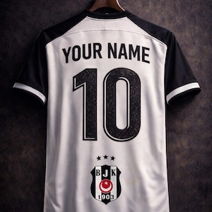 May include: White and black soccer jersey with the text "YOUR NAME" and the number "10" printed on the back. The jersey also features a team crest with a red crescent moon and star design. The jersey is hanging on a wooden hanger.
