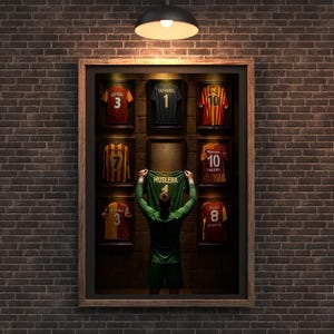 Pode incluir: Exibição emoldurada de camisas de futebol, incluindo uma camisa verde levantada por uma pessoa. As camisas são de várias cores, incluindo vermelho, amarelo e verde, com números e nomes visíveis. A exibição é colocada contra uma parede de tijolos.