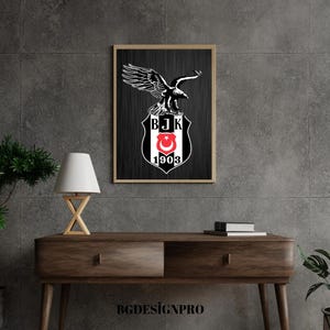 May include: Framed wall art featuring a black and white Besiktas JK emblem with an eagle and the text "BJK 1903". The artwork is displayed on a dark gray wall above a wooden console table with a lamp and books.