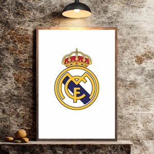 Puede incluir: Impresión enmarcada con el escudo del Real Madrid. El escudo es un círculo dorado con un diseño azul y blanco, coronado por una corona roja y dorada. La impresión se muestra en una estantería de madera contra una pared texturizada.