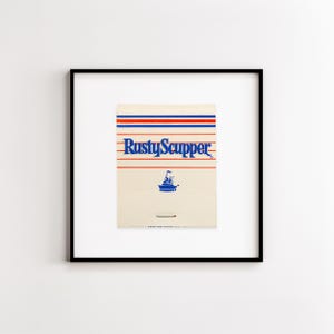May include: A square black frame containing a vintage matchbook cover with a cream background. The design features horizontal red and blue stripes, the blue text RustyScupper, and a small blue graphic of a boat. A single match is visible.