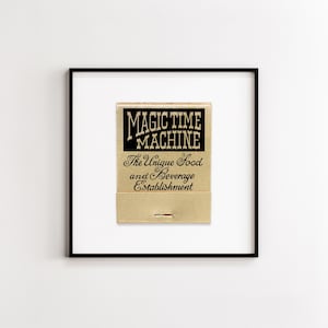 May include: A framed print featuring a gold-colored matchbook cover with black text that reads MAGIC TIME MACHINE The Unique Food and Beverage Establishment. The artwork is centered within a thin black frame against a plain white background.