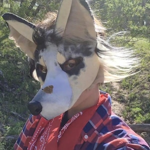 May include: A detailed close-up of a wolf-like mask with large, pointed ears. The mask features a white base with black and gray markings, a black nose, and visible eyes. The person is wearing a red bandana and a red and blue plaid shirt.