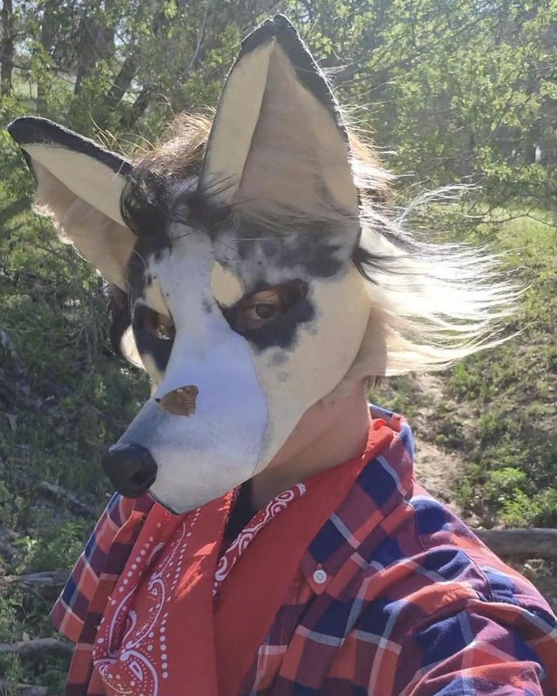 May include: A detailed close-up of a wolf-like mask with large, pointed ears. The mask features a white base with black and gray markings, a black nose, and visible eyes. The person is wearing a red bandana and a red and blue plaid shirt.