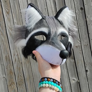 May include: A raccoon mask with a black, white, and gray color scheme. The mask has a black nose, eye openings, and ear tips. The ears and cheeks have white and gray fur. The mask is held up against a wooden background.
