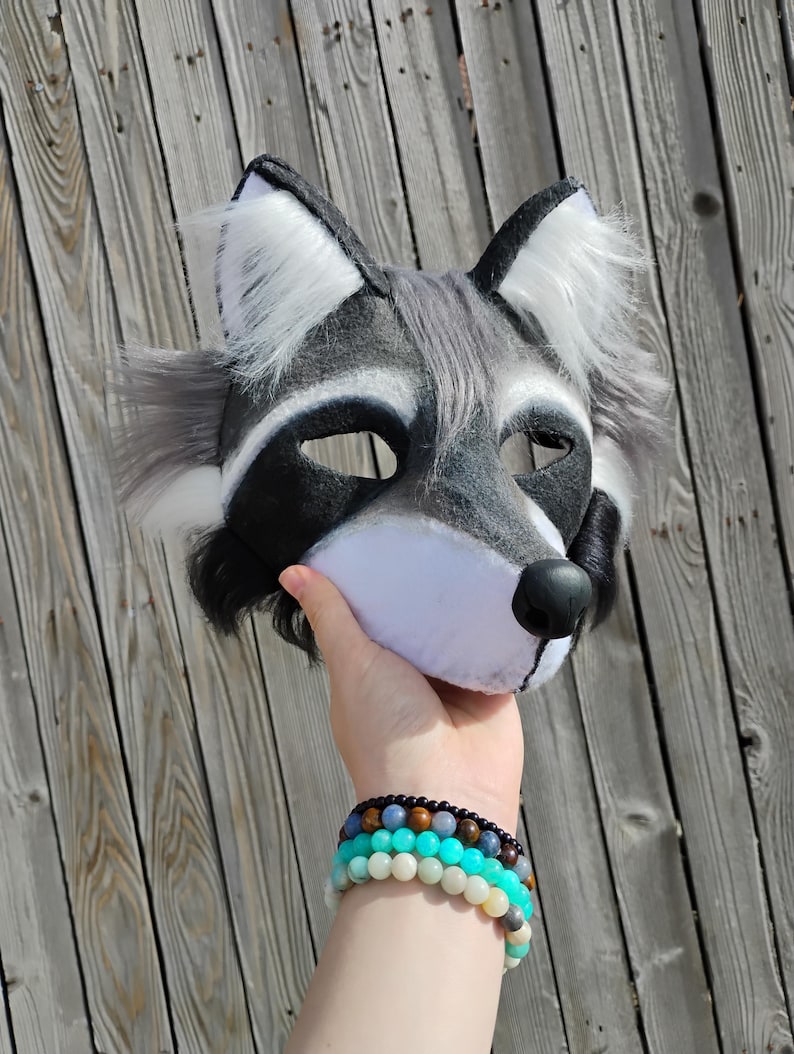 May include: A raccoon mask with a black, white, and gray color scheme. The mask has a black nose, eye openings, and ear tips. The ears and cheeks have white and gray fur. The mask is held up against a wooden background.