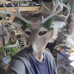 May include: A deer mask with white antlers and green leaf accents. The mask has a realistic deer face with brown and tan fur details. The person is wearing a gray and blue long-sleeved shirt.