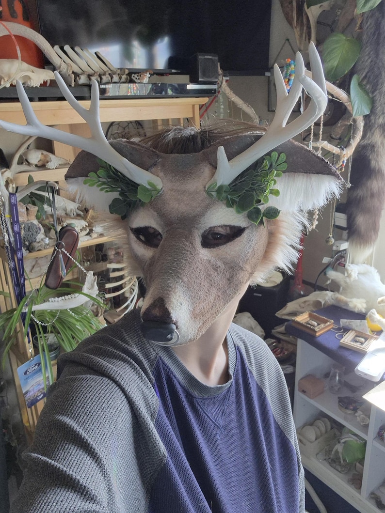 May include: A deer mask with white antlers and green leaf accents. The mask has a realistic deer face with brown and tan fur details. The person is wearing a gray and blue long-sleeved shirt.