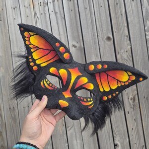 May include: A black felt mask with butterfly wing designs in orange and yellow. The mask has long, pointed ears and black fur accents. The mask is held up against a wooden background.