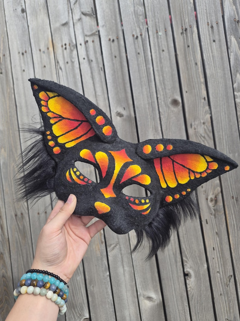 May include: A black felt mask with butterfly wing designs in orange and yellow. The mask has long, pointed ears and black fur accents. The mask is held up against a wooden background.