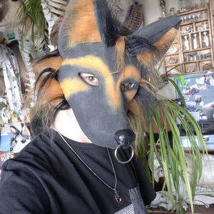 May include: A German Shepherd-style fursuit head with black, tan, and brown fur. The mask has a black nose and a silver ring. The person is wearing a black t-shirt with a geometric pattern on the bottom and a silver necklace.