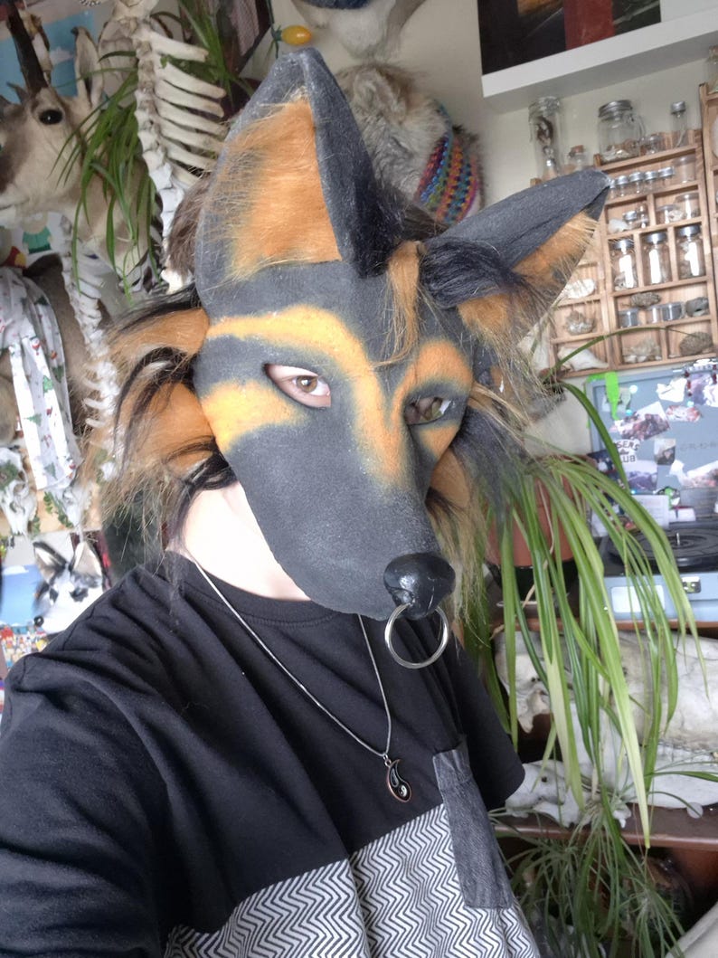 May include: A German Shepherd-style fursuit head with black, tan, and brown fur. The mask has a black nose and a silver ring. The person is wearing a black t-shirt with a geometric pattern on the bottom and a silver necklace.