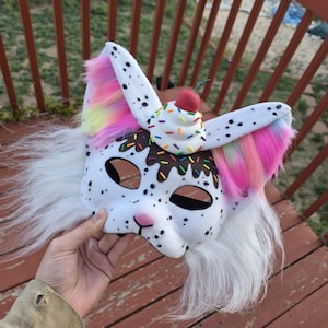 May include: A white and black spotted mask with a cupcake design on top. The mask has long white fur and colorful accents on the ears. The cupcake has sprinkles and a cherry on top.