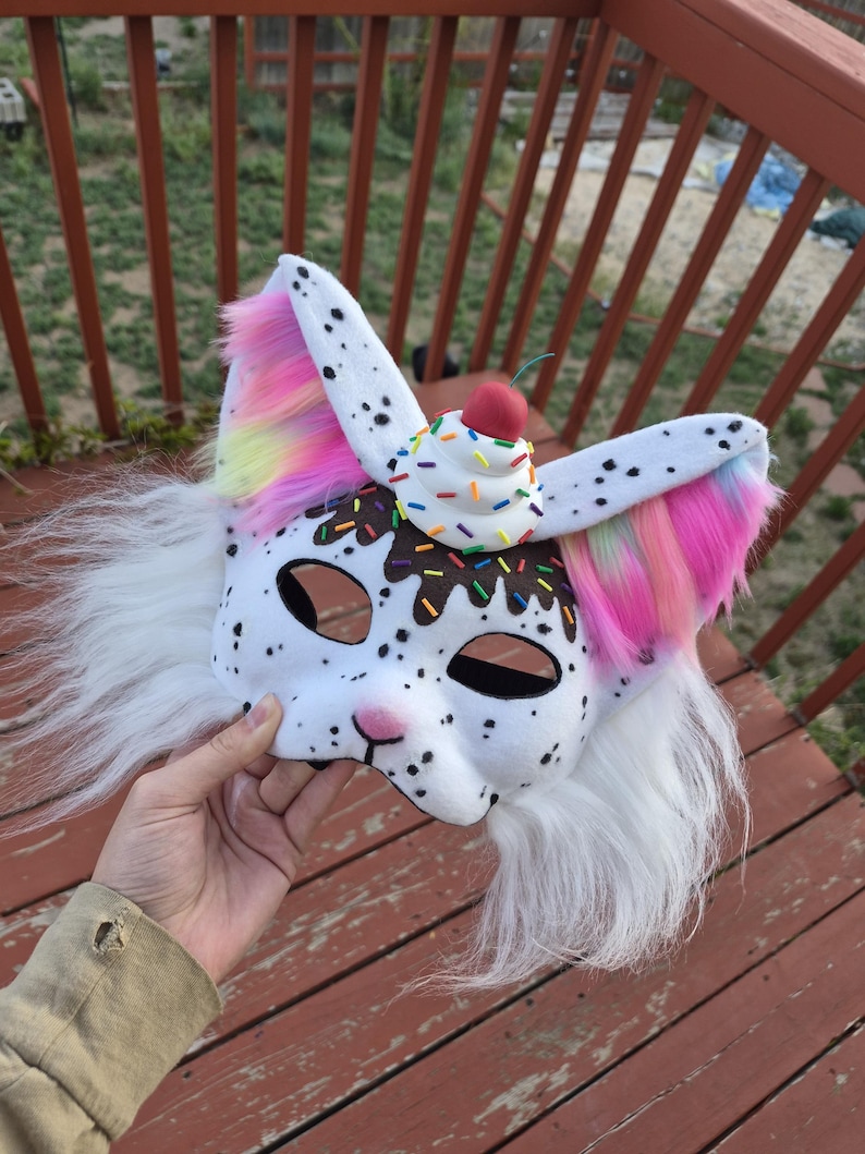 May include: A white and black spotted mask with a cupcake design on top. The mask has long white fur and colorful accents on the ears. The cupcake has sprinkles and a cherry on top.