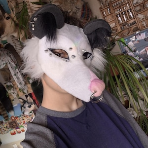 May include: A white and gray possum mask with black ears and silver studs. The mask has a pink nose and a silver ring. The person is wearing a blue and gray long-sleeve shirt.