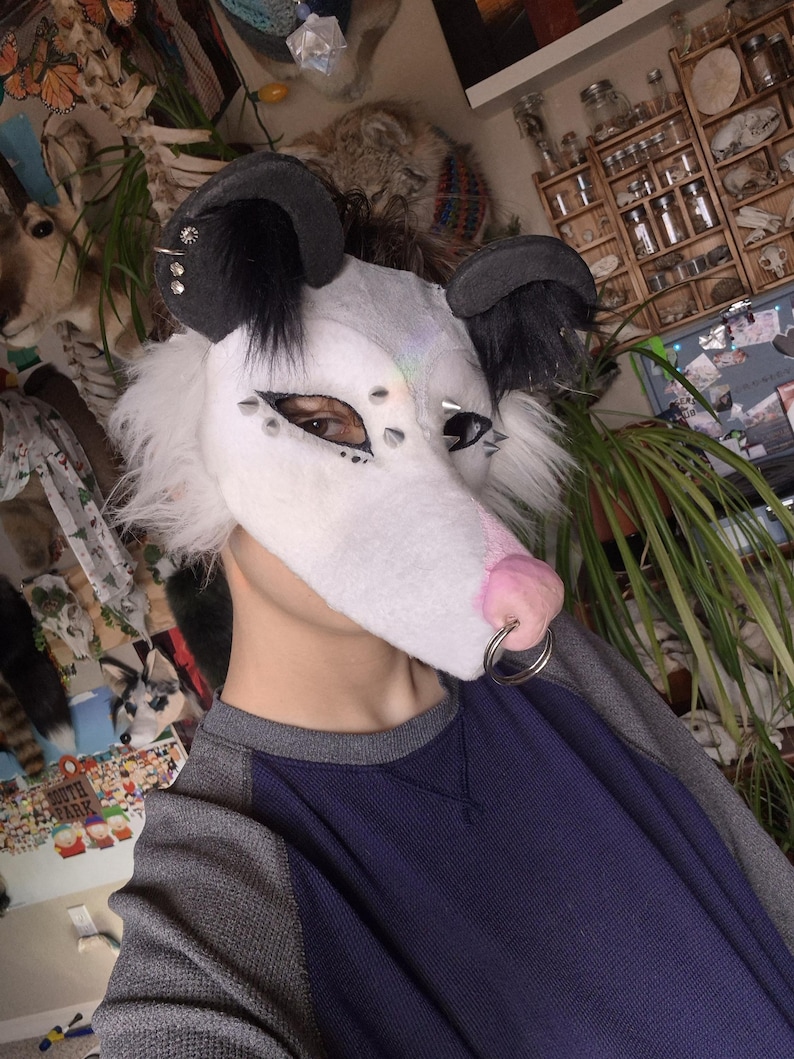 May include: A white and gray possum mask with black ears and silver studs. The mask has a pink nose and a silver ring. The person is wearing a blue and gray long-sleeve shirt.