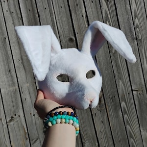 May include: A white bunny mask with long, upright ears. The mask has eye openings and a pink nose. The mask is held up against a weathered wooden background. The mask is made of a soft, plush material.