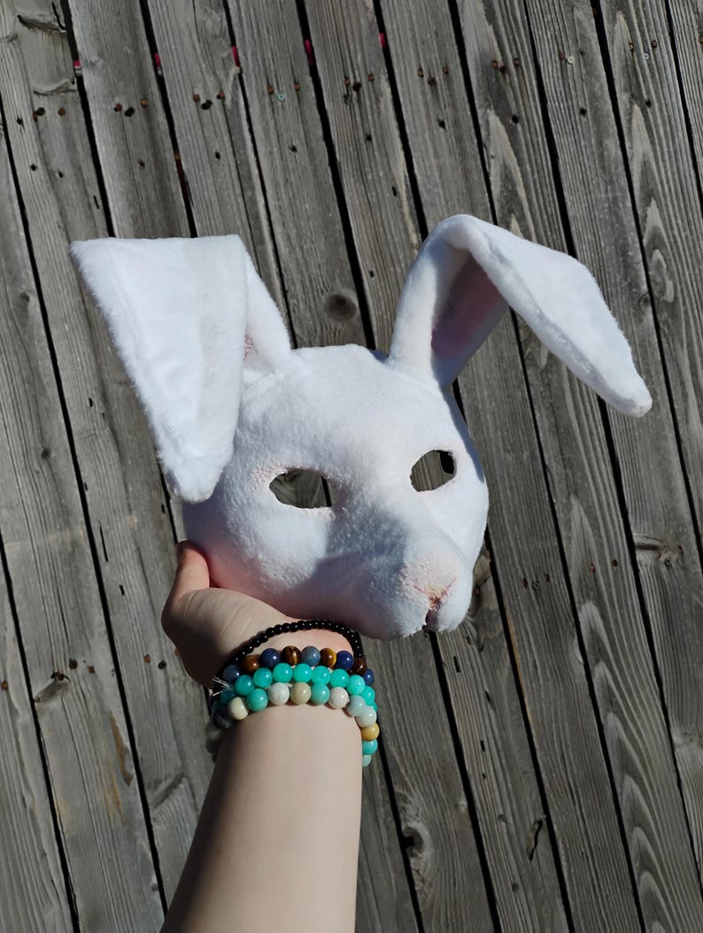 May include: A white bunny mask with long, upright ears. The mask has eye openings and a pink nose. The mask is held up against a weathered wooden background. The mask is made of a soft, plush material.