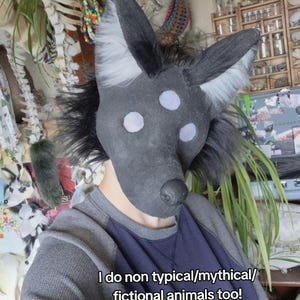 May include: A dark gray and black animal mask with three white eye spots and large ears. The mask has a black nose and is surrounded by black fur. The text on the image reads, "I do non typical/mythical/fictional animals too!"