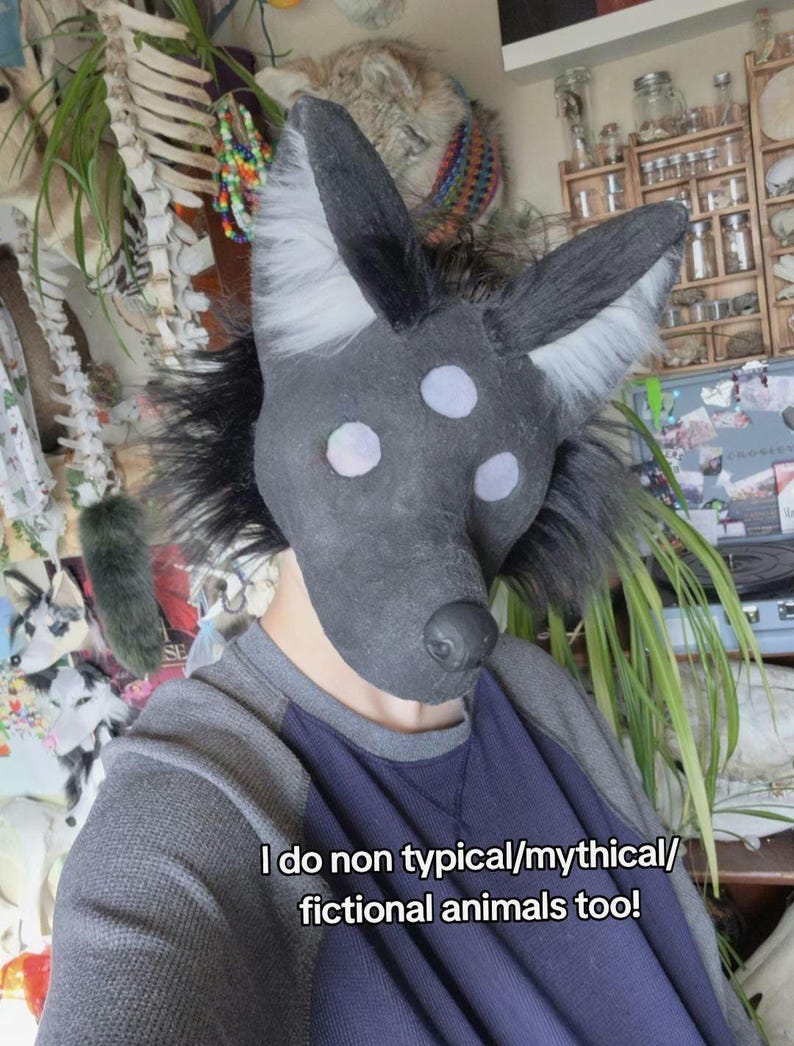 May include: A dark gray and black animal mask with three white eye spots and large ears. The mask has a black nose and is surrounded by black fur. The text on the image reads, "I do non typical/mythical/fictional animals too!"