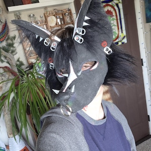 May include: A black and gray furry mask with large pointed ears, adorned with silver accents and bottle cap details. The mask features a white star design on the forehead and a silver nose ring. The person is wearing a gray and blue shirt.