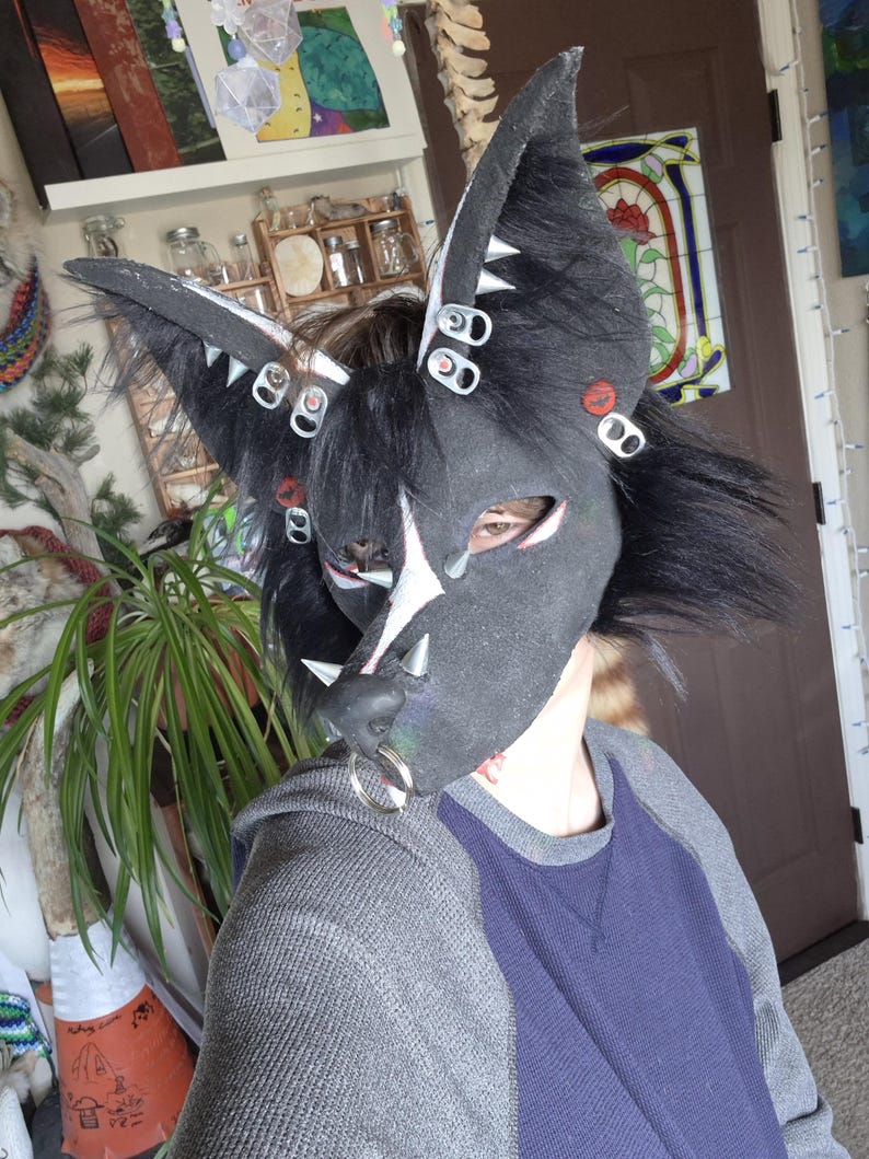 May include: A black and gray furry mask with large pointed ears, adorned with silver accents and bottle cap details. The mask features a white star design on the forehead and a silver nose ring. The person is wearing a gray and blue shirt.