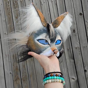 May include: A handmade animal mask with a wolf-like design. The mask features blue eyes, a tan nose, and white and gray fur. The ears are brown with white fur accents. The mask is held up against a wooden background.