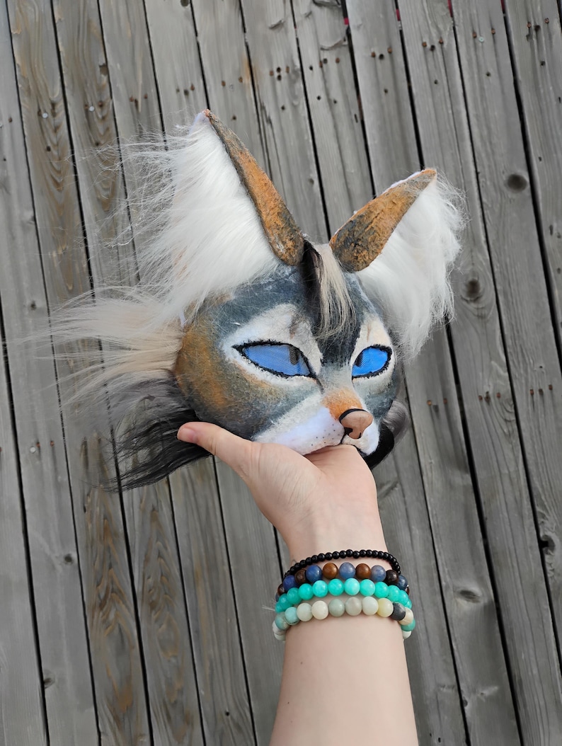 May include: A handmade animal mask with a wolf-like design. The mask features blue eyes, a tan nose, and white and gray fur. The ears are brown with white fur accents. The mask is held up against a wooden background.
