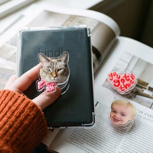 Può includere: Un supporto per telefono trasparente con un gatto eterocromatico che indossa un fiocco rosa a pois, fissato su un e-reader nero. Sono visibili altri due supporti: uno a forma di cuore con il testo rosso you got this e uno con il volto di un bambino.