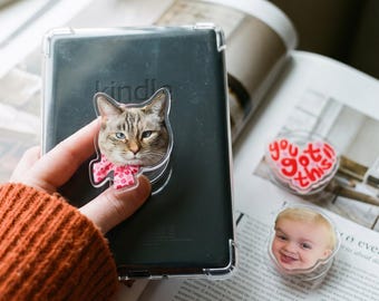 May include: A clear phone grip featuring a cat with heterochromia wearing a pink polka dot bow, attached to a black e-reader. Two additional grips are visible nearby: one heart-shaped with the red text you got this and one with a child's face.