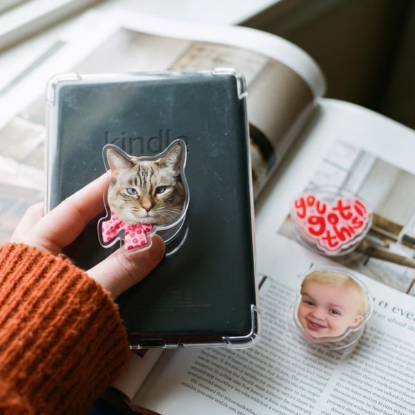 May include: A clear phone grip featuring a cat with heterochromia wearing a pink polka dot bow, attached to a black e-reader. Two additional grips are visible nearby: one heart-shaped with the red text you got this and one with a child's face.