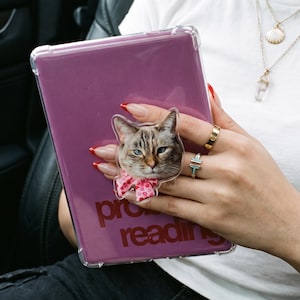 Può includere: Una custodia trasparente per tablet con un supporto per telefono a forma di gatto, caratterizzato da un muso di gatto realistico e un fiocco rosa a fantasia. Il tablet è viola e riporta la scritta "pro reading" in rosso scuro.