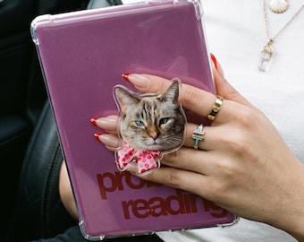 May include: A clear tablet case featuring a cat-shaped phone grip with a realistic cat face and a pink patterned bow. The tablet case is purple and displays the text "pro reading" in a dark red font, providing a clear view of the device.