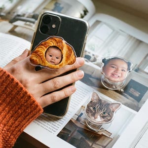 May include: Three circular phone grips featuring custom photographic prints. One grip shows a baby's face inside a golden-brown croissant, another shows a smiling child's face, and the third displays the face of a brown and gray tabby cat.