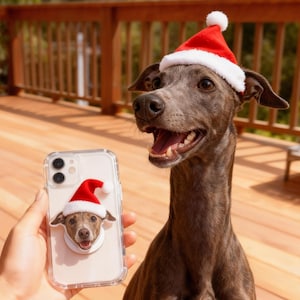 May include: A grey dog wearing a red and white Santa hat sits on a wooden deck. A clear phone case with a matching dog wearing a Santa hat is held in front of the dog. The dog is smiling with its mouth open.