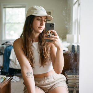 May include: A beige baseball cap with a small embroidered design on the front, worn with a white sleeveless crop top and beige shorts. A smartphone with a dog-themed pop socket is held up, and a small cherub tattoo is visible on the forearm.