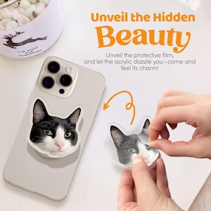 May include: A white phone case with a black and white cat sticker on it. The sticker is being peeled off to reveal a clear acrylic case underneath. The text "Unveil the Hidden Beauty" is written in orange letters on a white background.