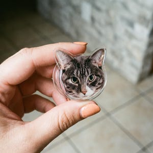 May include: A circular phone grip featuring a high-resolution print of a brown and grey tabby cat face with green eyes and a white muzzle. The accessory has a clear, cat-ear-shaped acrylic border and a collapsible plastic base for device support.