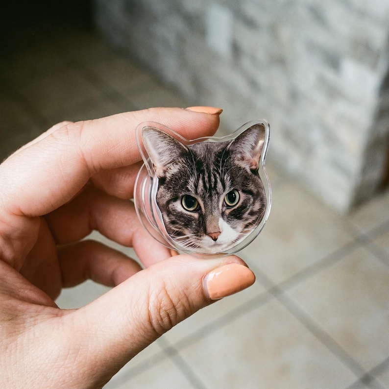 May include: A circular phone grip featuring a high-resolution print of a brown and grey tabby cat face with green eyes and a white muzzle. The accessory has a clear, cat-ear-shaped acrylic border and a collapsible plastic base for device support.
