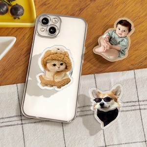 May include: Three phone stickers featuring a cat wearing a brown fuzzy hat, a baby wearing a light blue shirt, and a dog wearing sunglasses and a black scarf.