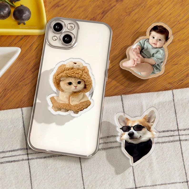 May include: Three phone stickers featuring a cat wearing a brown fuzzy hat, a baby wearing a light blue shirt, and a dog wearing sunglasses and a black scarf.