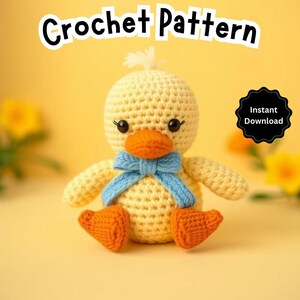 May include: A crocheted yellow duck with a blue bow, sitting on a yellow surface. The duck has black eyes and an orange beak. The text "Crochet Pattern" is at the top of the image, and "Instant Download" is in a black circle at the top right.