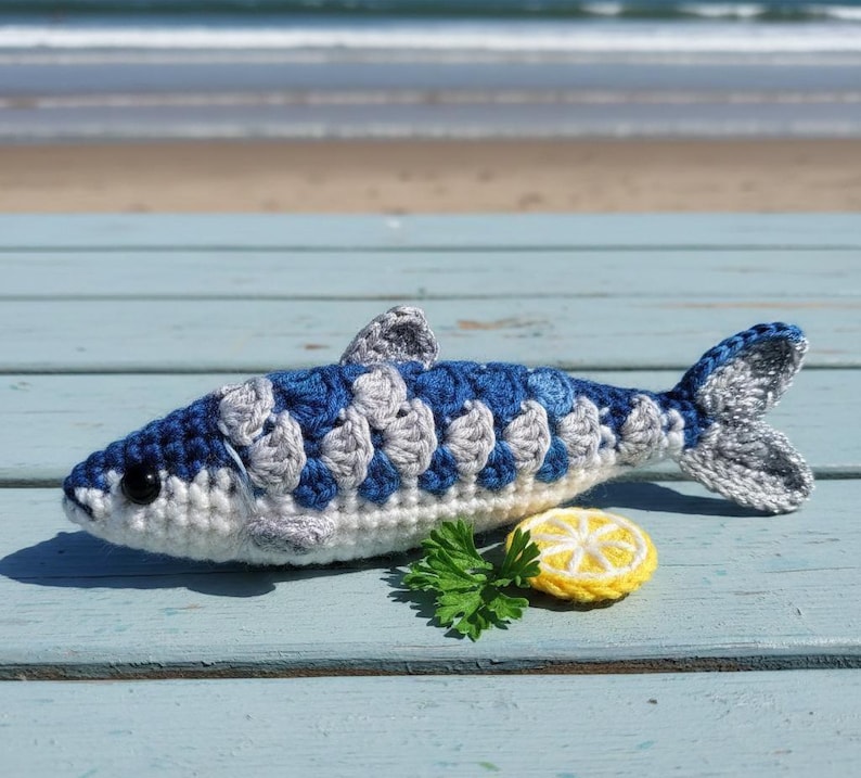 May include: A crocheted fish in shades of blue, gray, and white, with a yellow lemon slice and green parsley. The fish has a detailed scale pattern and a fin. The scene is set on a light blue wooden surface, with the ocean in the background.