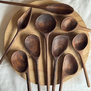 Può includere: Una collezione di cucchiai di legno fatti a mano in varie forme e dimensioni, disposti su un vassoio rotondo di legno. I cucchiai sono di un ricco colore marrone scuro, che mostra la venatura naturale del legno. Lo sfondo è un tessuto bianco.