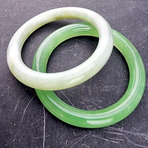 May include: Two jade bangles. One is a pale green color, and the other is a vibrant green. The bangles are circular and smooth, with a polished finish. They are displayed on a dark, textured surface.