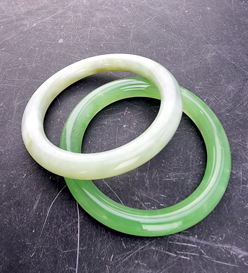 May include: Two jade bangles. One is a pale green color, and the other is a vibrant green. The bangles are circular and smooth, with a polished finish. They are displayed on a dark, textured surface.