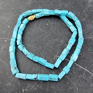 Puede incluir: Un collar compuesto por cuentas rectangulares de color turquesa con vetas oscuras. Las cuentas están ensartadas con un cierre dorado. El collar se exhibe sobre una superficie oscura.