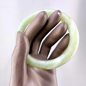 May include: A pale green jade bangle bracelet. The smooth, circular bracelet is held by a hand. The jade has a translucent quality, with variations in color. The bracelet is a classic piece of jewelry.